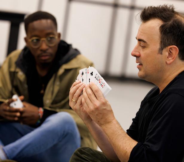 Steve Cuiffo穿着海军蓝衬衫，手里拿着一副扑克牌在演示. 一个学生坐在地板上，专心地观察着. 设置是非正式的和教育的，暗示动手学习的经验.